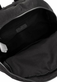 Wnętrze czarnego plecaka z gładką podszewką wykonaną z tkaniny, wyposażone w zapinaną na zamek kieszeń oraz metkę marki. Wnętrze jest przestronne i pozbawione ozdób.