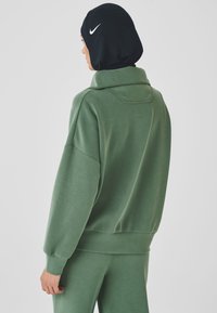 Green sweatshirt with a high collar and dropped shoulders, made of soft fabric. Features a black hijab with a white Nike logo accent.