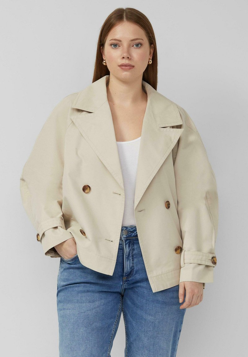 Frau trägt eine beige zweireihige Jacke über einem weißen Oberteil und blauen Jeans, steht mit einer Hand in der Tasche vor einem grauen Hintergrund.