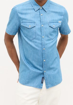 Hombre con camisa de mezclilla azul claro de manga corta con bolsillos en el pecho abotonados y pantalones blancos, con las manos detrás de la espalda.