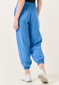 Blauwe, lichte, elastische broeken met gewatteerde boorden. Gladde textuur, casual pasvorm, gestyled met witte sportschoenen.