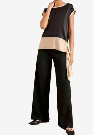 Femme portant un haut noir et beige à manches courtes avec un détail nœud sur le côté et un pantalon noir à jambes larges, associé à des ballerines pointues noires.