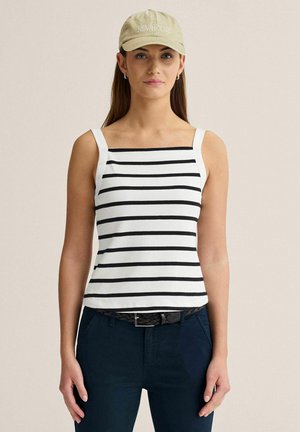 Kvinde iført en beige kasket, sort-hvid stribet ærmeløs top, mørke bukser med et fletbælte i sort, stående foran en ensfarvet baggrund.