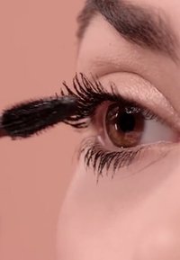 Black mascara brush applying product on long, dark eyelashes. The eye is brown, with a soft pink background highlighting the features.