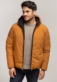 Brązowa kurtka puchowa z czarnym wnętrzem, z kapturem i zamkiem błyskawicznym z przodu. Model ma na sobie jasnoszary sweter pod spodem.