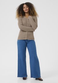 Beige knitted cardigan with button closure and textured sleeves, paired with high-waisted blue flared jeans and dark shoes.