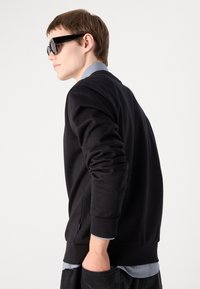 Sweatshirt preto de mangas longas, gola redonda, com detalhes em zíper lateral, sobre uma camisa azul clara em camadas, combinado com jeans escuros.