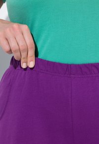 Paarse elastische taillebandbroek met een zachte textuur, gecombineerd met een groene top met korte mouwen, waarbij een close-up van het detail van de tailleband wordt weergegeven.