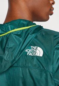 Groene lichtgewicht jack met een capuchon, voorzien van een gestructureerde stof, gele accenten langs de naad, en een witte logoafdruk op de achterkant.