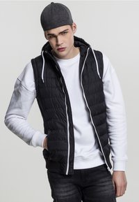 Vest puf din matlasat negru cu fermoar alb, purtat peste un hanorac alb. Modelul poartă o pălărie gri închis.