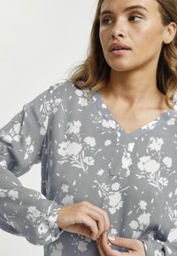 Blusa floreale grigia con motivi di fiori bianchi, maniche lunghe, scollatura a V e una fine trama a righe. Dettaglio con bottoni ai polsi.