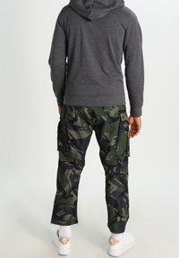 Pantalones cargo de camuflaje en verde y negro, con múltiples bolsillos. Llevados con una sudadera gris y zapatillas blancas, vistos desde atrás.