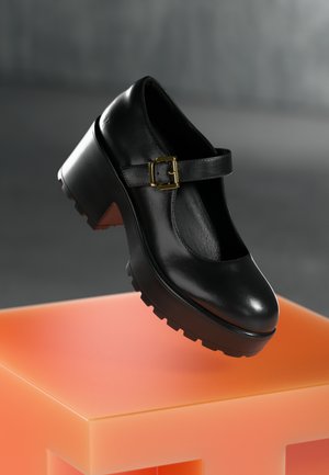 Black leather shoe with a glossy finish, chunky heel, buckle strap, and rugged outsole, set on a vibrant orange block.