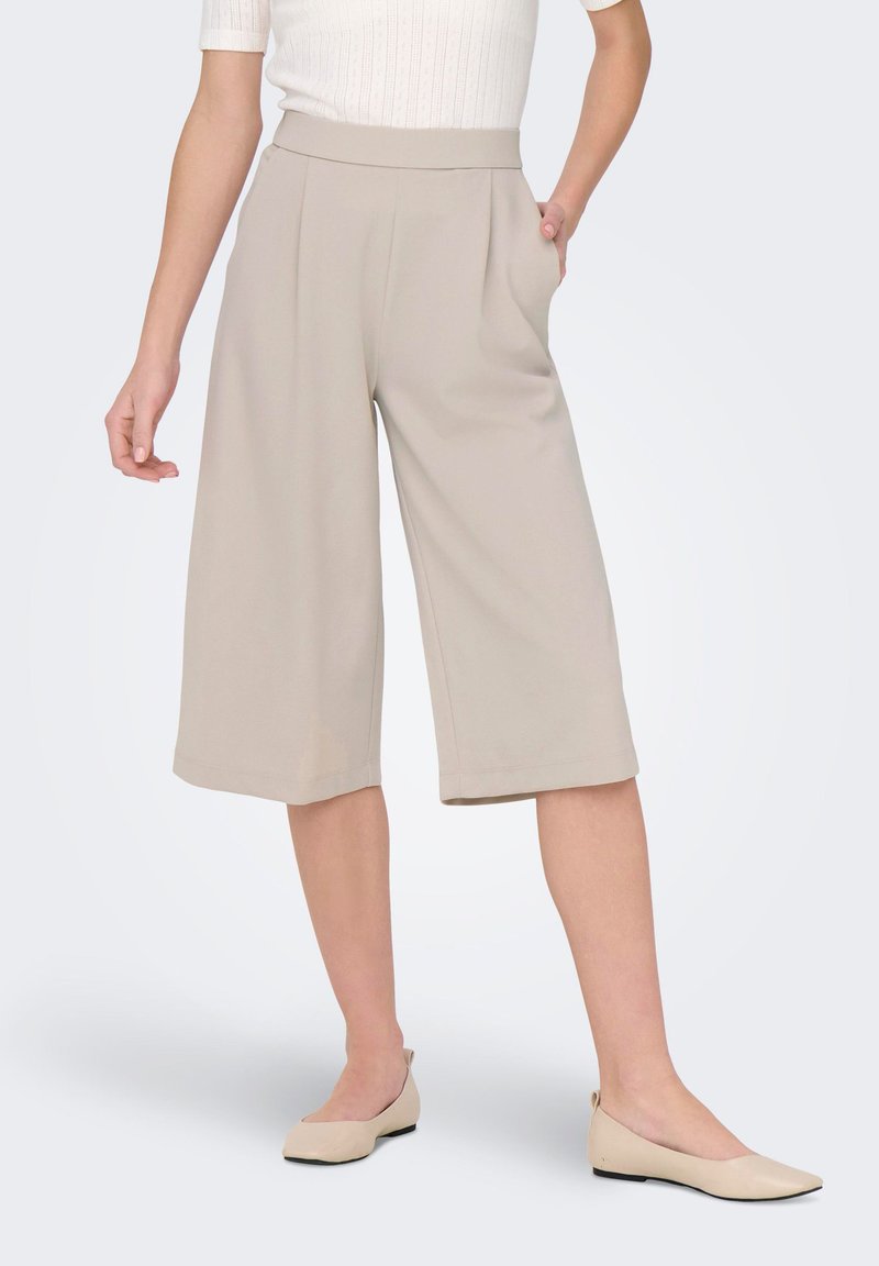 Kvinde iført beige vide kortere bukser og beige flade sko, med den ene hånd i lommen, mod en ensartet lys baggrund.