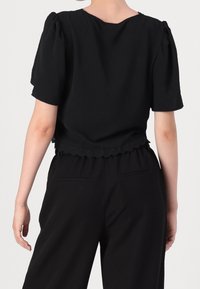 Svart kortärmad topp med rundad halsringning och draperad axeldetalj; fållen har vågigt avslut; tyget verkar lätt och mjukt.