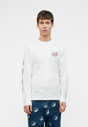 Jauns vīrietis ar baltu garām piedurknēm kreklu, kurā ir krāsaini piedurkņu printi un "Santa Cruz" logo, kombinēts ar ziliem džinsiem ar jin-janga simboliem.