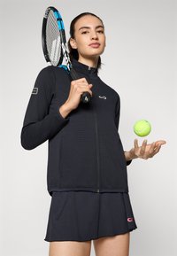 Svart zip-up treningsjakke med teksturert stoff, kombinert med et mørkt skjørt. Holder en tennisracket og en lys gul tennisball.