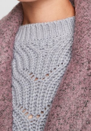 Suéter gris de punto con patrón texturizado y puntos abiertos, llevado debajo de una prenda exterior peluda de color rosa con un diseño moteado.