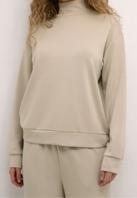 Sweat-shirt beige avec un col montant, des manches longues et des poignets côtelés. Tissu lisse avec une coupe décontractée et un bord inférieur à bandes.