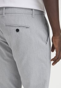Pantaloni grigio chiaro realizzati in un tessuto liscio, con una tasca laterale e una tasca posteriore con bottone nero e fodera interna a righe.