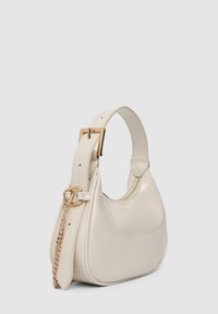 Sac à main beige breveté avec un design incurvé, une finition brillante, une quincaillerie dorée et une bandoulière ornée d'une chaîne. Dispose d'une fermeture éclair.