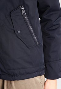 Tummansininen nailonitakki, jossa on vetoketjullinen tasku, läppätasku sekä joustavat hihansuut ja tikattu pinta helmassa. Kestävä ja toiminnallinen muotoilu.