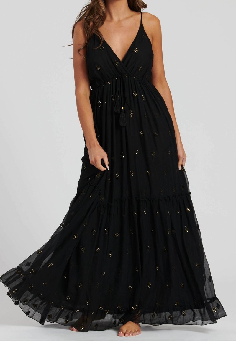 Femme portant une longue robe noire sans manches avec un motif à pois dorés, tenant l'ourlet, pieds nus, sur un fond gris uni.