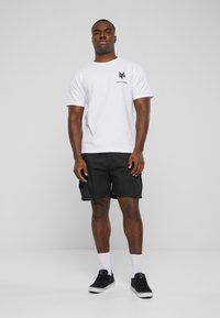 Hvid T-shirt med korte ærmer og sort "Zoo York" logo, parret med sorte cargo shorts. Modellen står med sorte sneakers og hvide sokker.