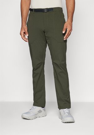 Man draagt olijfgroene broek met zwarte verstelbare riem en witte sportschoenen, staand tegen een effen lichte achtergrond.