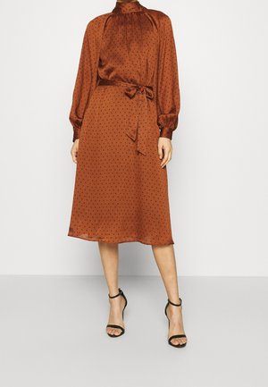 Kobieta ubrana w rdzawobrązową, midi sukienkę z długim rękawem i czarnymi kropkami, związaną w talii, oraz czarne sandały na kostkę, stojąca na szarej podłodze.