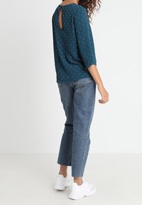 Mujer vestida con blusa azul estampada con escote en forma de llave, jeans capri de cintura alta y zapatillas blancas, de pie contra un fondo blanco.
