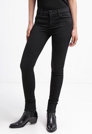 Kobieta ubrana w czarne obcisłe jeansy i czarne botki, stojąca na jednolitym białym tle.