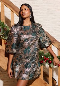 Kleid aus metallischem Stoff mit grünem Blumenmuster auf bronzefarbenem Hintergrund. Kurze Ärmel und ein geraffter Saum.