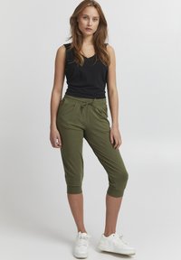 Kvinne iført svart ermeløs topp, olivengrønne, cropped bukser med snøring, og hvite joggesko, som står mot en ensfarget lys bakgrunn.