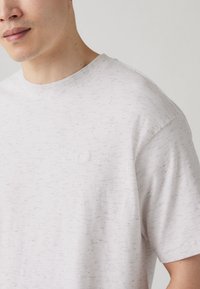 Light gray t-shirt made of textured fabric, featuring a crew neck and short sleeves. Small embossed logo on the chest.