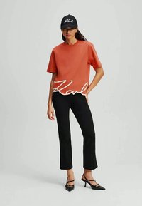 T-shirt in cotone arancione con logo bianco "Karl", abbinata a pantaloni aderenti neri e décolleté nere con punta appuntita. Modella che indossa un cappellino nero coordinato.