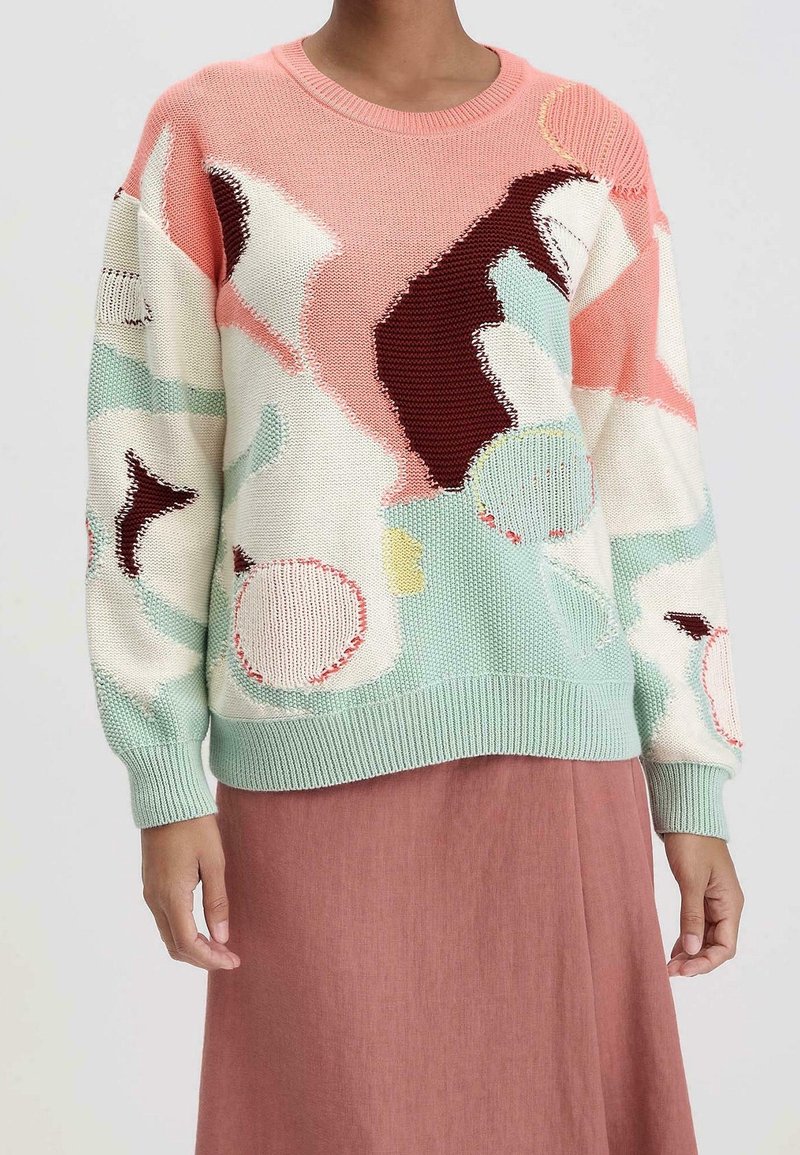 Personne portant un pull en tricot à manches longues avec des formes abstraites pastel en rose, crème, menthe et bordeaux, associé à une jupe rose atténuée.