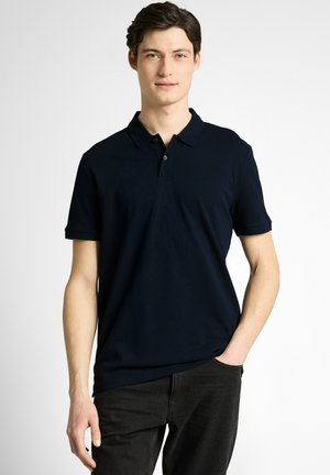 Młody mężczyzna o ciemnych włosach, ubrany w czarną, krótkorękawową koszulkę polo i czarne spodnie, stojący z jedną ręką w kieszeni na białym tle.