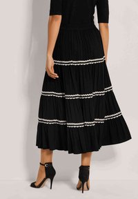 Schwarzer, gestufter Rock mit weißen, gewellten Besatzdetails, aus leichtem Stoff gefertigt. Kombiniert mit Sandalen mit Knöchelriemen, von hinten präsentiert.