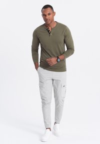 Olivgrünes Langarm-Henley-Shirt mit Dreiknopfverschluss, kombiniert mit hellgrauen Cargo-Hosen mit Seitentaschen und weißen Sneakers.