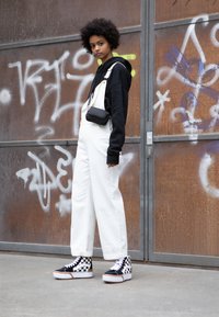 Overoles blancos de pierna ancha, sudadera negra y zapatillas altas a rayas en blanco y negro. Pequeño bolso negro cruzado colgado del hombro.