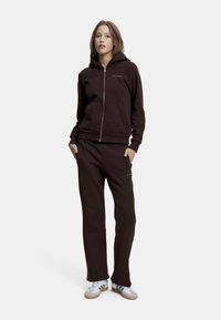 Brun zip-up hettegenser med kengurulommer og logo. Kombinert med matchende joggebukser, begge laget av myk, teksturert stoff, og brukt med hvite joggesko.