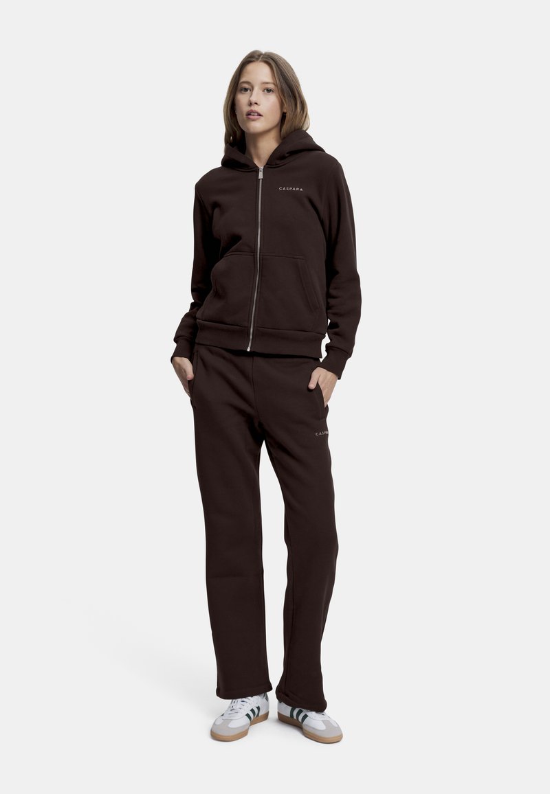 Brun zip-up hettegenser med kengurulommer og logo. Kombinert med matchende joggebukser, begge laget av myk, teksturert stoff, og brukt med hvite joggesko.