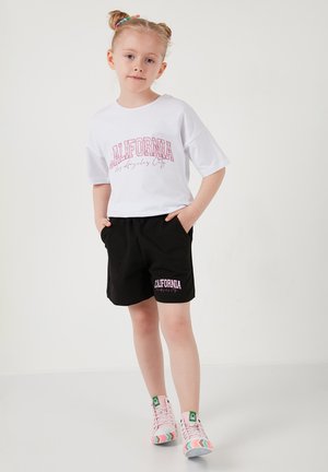 Ung pige med to knolde, iført hvid "California" t-shirt, sorte shorts med "California"-tryk og lyserøde sneakers med mønster, står mod en hvid baggrund.