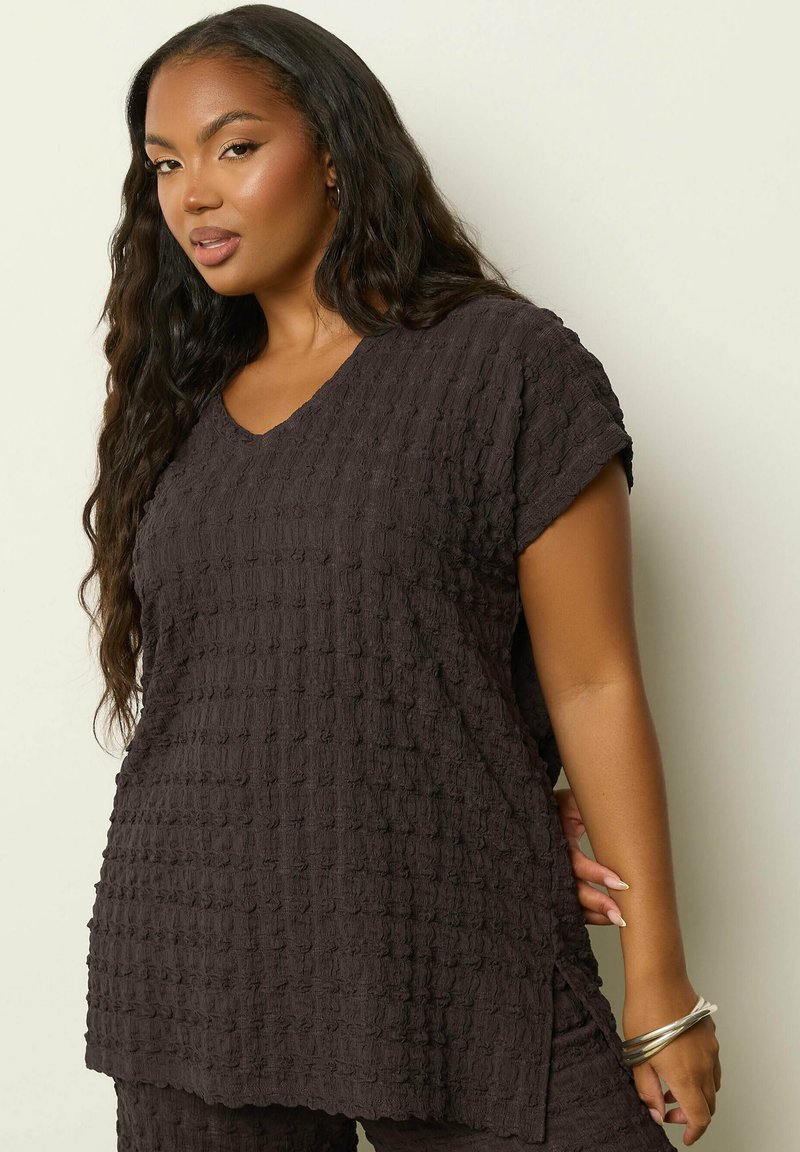 Nainen, jolla on pitkät, laineikkaat hiukset, pukeutuneena teksturoituun mustaan lyhythihaiseen toppiin ja siihen sopiviin housuihin, hopeiset rannelyönnit ranteessaan, yksinkertaisella taustalla.