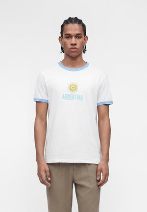 Νέος άνδρας με πλεξούδες, φορώντας ένα λευκό μπλουζάκι με ανοιχτό μπλε περίγραμμα, ένα γραφικό ήλιου και το κείμενο "ARGENTINA", στέκεται μπροστά σε ένα απλό φόντο.