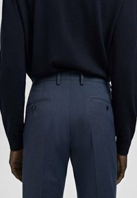 Tył osoby ubranej w dopasowane, niebieskie spodnie garniturowe z zapinanymi na guziki tylnymi kieszeniami oraz w czarną, włożoną do środka koszulę z długim rękawem.