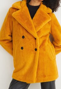 Cappotto giallo peloso a doppio petto con ampio risvolto e bottoni neri, dotato di due tasche laterali, morbido al tatto e dalla vestibilità rilassata.