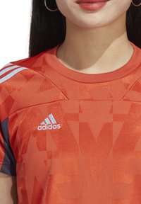 T-shirt de sport rouge avec un motif géométrique, fabriqué dans un tissu lisse, avec le logo Adidas blanc et trois bandes blanches sur l'épaule.