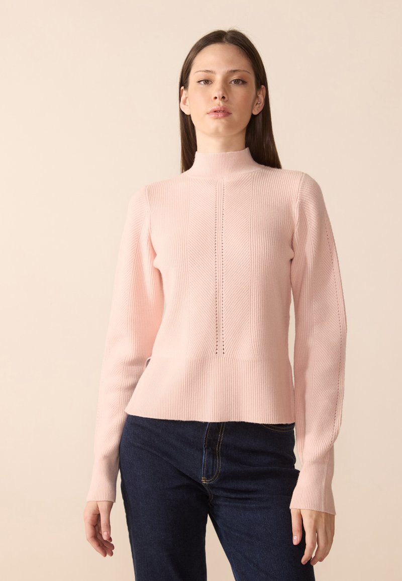 Suéter suave em rosa claro, com gola alta e detalhes verticais intrincados. Mangas longas com punhos ajustados, combinado com jeans azul escuro.
